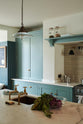 A spacious yet modest Trinity Blue classic English kitchen features a Silestone worktop sink run with a single fluted Belfast sink and aged brass Ionian tap, glazed cupboards, and a kitchen island with a copper sink—all topped off with a white porcelain splashback. Lovingly decorated with a silver juicer, papayas, live basil, berries, dried ingredients in glass jars, the room feels humble, warm, and inviting.