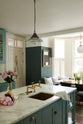A close, angled view of a spacious yet modest Trinity Blue classic English kitchen features a marble worktop island with a copper sink and aged brass Mayan taps, alongside a breakfast table by the arched window. Lovingly decorated with pink peonies in a vase, glass pendant lights, and a small blackboard and book, the room feels humble, warm, and inviting.