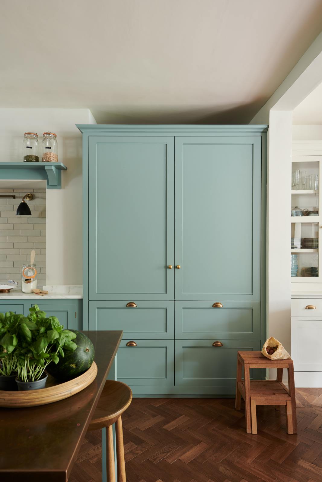 A spacious yet modest Trinity Blue classic English kitchen features a Silestone worktop cooker run, an overmantle canopy with a white porcelain splashback, a large full-height cabinet housing a fridge/freezer, a copper prep table, and deVOL wooden bum stools. Lovingly decorated with a watermelon, live basil, and dried ingredients in glass jars, the room feels humble, warm, and inviting.