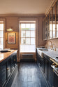Nestled between the sink and cooker run of a bold and dramatic Haberdasher’s kitchen, which features an Arabescato worktop, a single fluted sink, an aged brass Ionian tap, a glazed wall cupboard, and a kitchen island with a copper worktop, is a bright view of Bath.