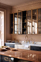 An oblique view of a bold and dramatic Haberdasher’s kitchen with a sink and cooker run, an Arabescato worktop and single fluted sink, an aged brass Ionian tap, a glazed wall cupboard, and a kitchen island with a copper worktop. Adorned with artisan ceramics, labelled glass jars, and a colourful sandwich, creating a local bristo bar in a Manhattan space.