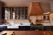A bold and dramatic Haberdasher’s kitchen with a sink and cooker run, an Arabescato worktop and single fluted sink, an aged brass Ionian tap, a glazed wall cupboard, a large copper canopy, and a kitchen island with a copper worktop. Adorned with copper pans, artisan ceramics, labelled glass jars, and a quirky framed illustration, creating a local bristo bar in a Manhattan space.