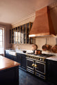 An oblique view of a bold and dramatic Haberdasher’s kitchen with a sink and cooker run, an Arabescato worktop and single fluted sink, an aged brass Ionian tap, a glazed wall cupboard, a large copper canopy, and a kitchen island with a copper worktop. Adorned with copper pans, artisan ceramics, aged brass hanging rails, and labelled glass jars, creating a local bristo bar in a Manhattan space.