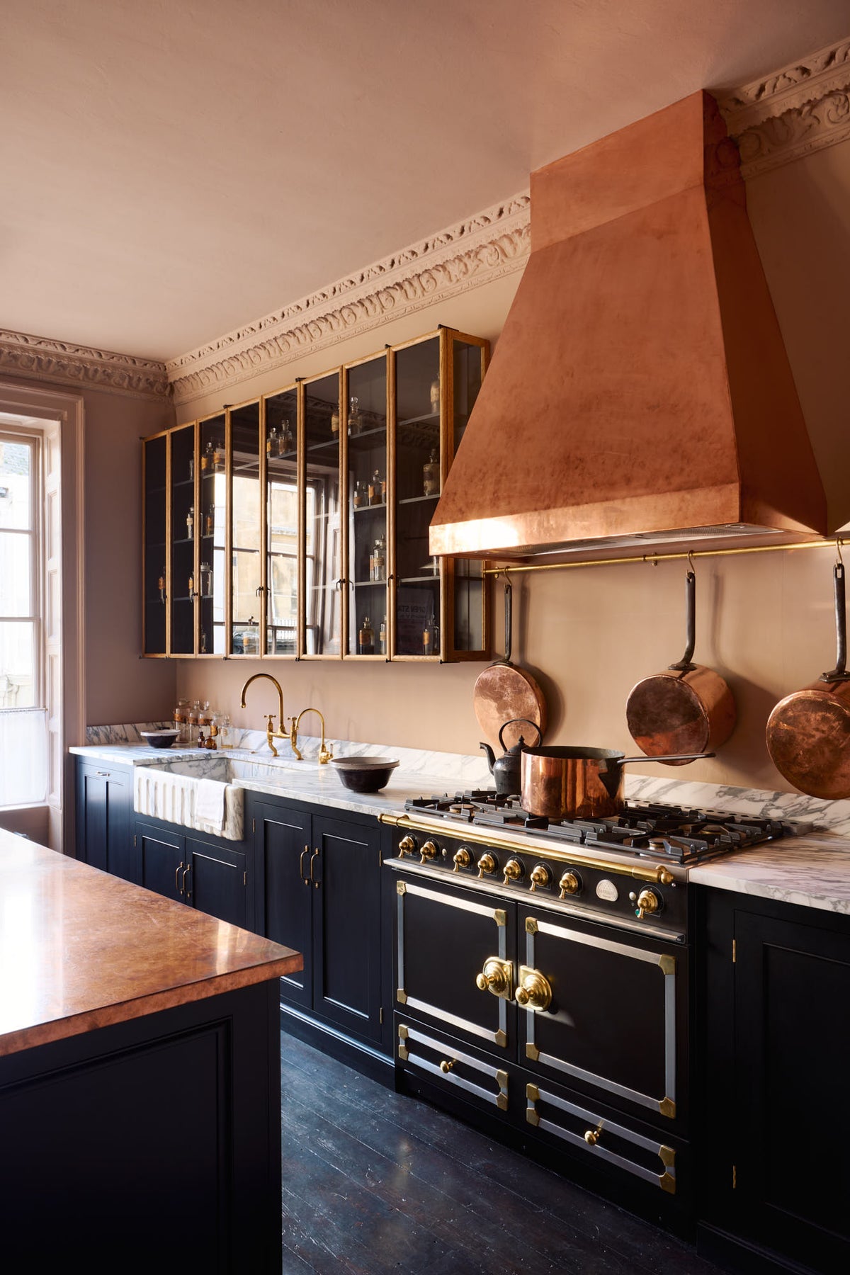 An oblique view of a bold and dramatic Haberdasher’s kitchen with a sink and cooker run, an Arabescato worktop and single fluted sink, an aged brass Ionian tap, a glazed wall cupboard, a large copper canopy, and a kitchen island with a copper worktop. Adorned with copper pans, artisan ceramics, aged brass hanging rails, and labelled glass jars, creating a local bristo bar in a Manhattan space.