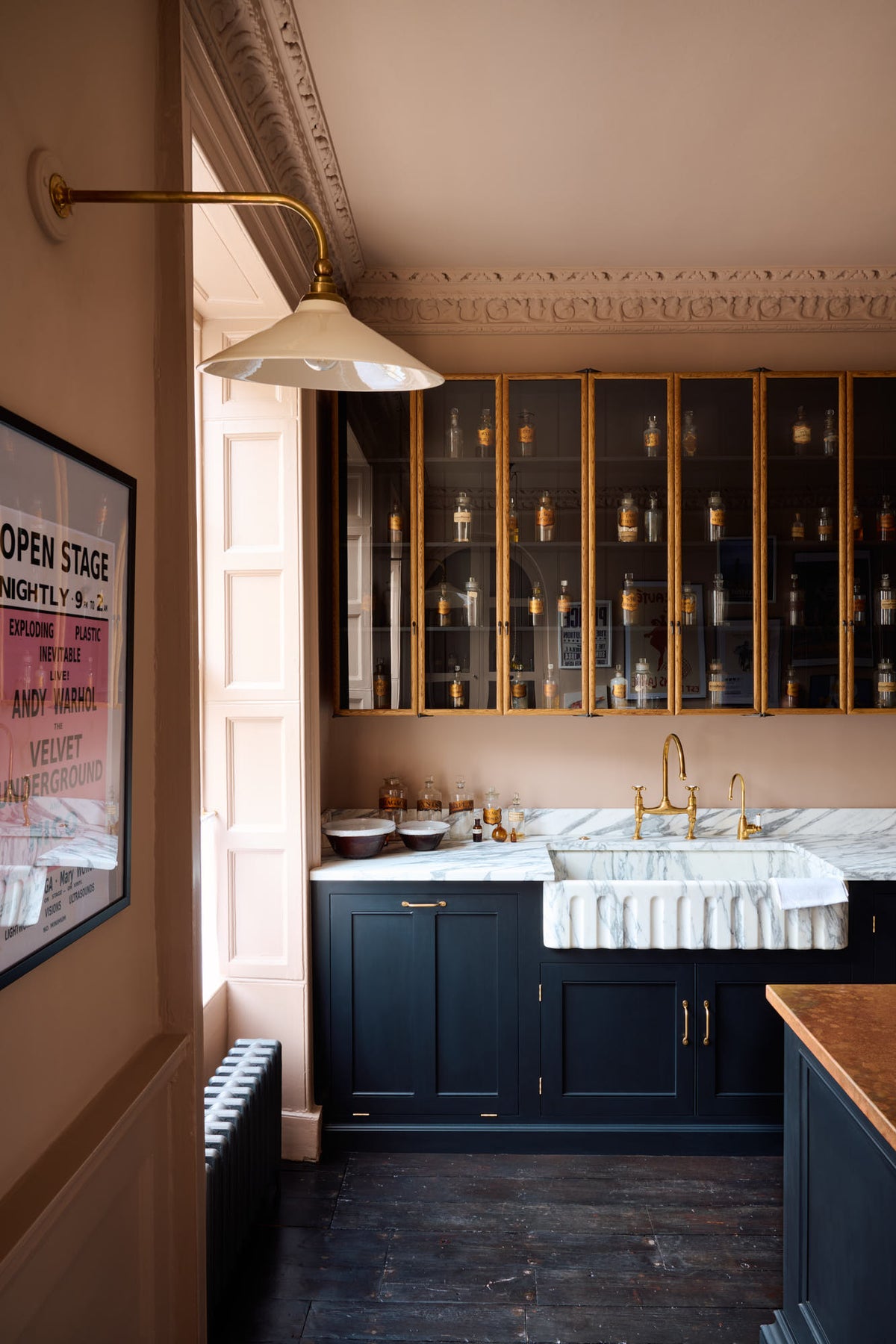 A quiet corner of a bold and dramatic Haberdasher’s kitchen with a sink and cooker run, an Arabescato worktop and single fluted sink, an aged brass Ionian tap, a glazed wall cupboard, and a kitchen island with a copper worktop. Adorned with a milk glass task light, artisan ceramics, labelled glass jars, and a framed poster, creating a local bristo bar in Manhattan space.