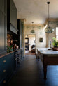 An elegant classic kitchen includes a cooker and sink with an aged copper worktop, a shaker canopy, counter cabinets, a marble worktop heirloom dairy table, a breakfast nook, and tall black chairs. The tall-ceilinged room has a rustic wash finish, with green draping down the walls, resembling overgrown ancient ruins. Beyond the archways leading to the living room is a striking black fireplace, a black-framed mirror, and a woven chandelier.