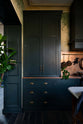 A closer look at an elegant classic kitchen’s cooker run reveals an aged copper worktop, a shaker canopy, counter cabinets, and a marble worktop heirloom dairy table. The pantry-blue counter cabinets stand tall, and beside them is a black gas cooker, copper pans, and fresh chillies hanging from aged brass rails, with a large aged copper splashback behind.