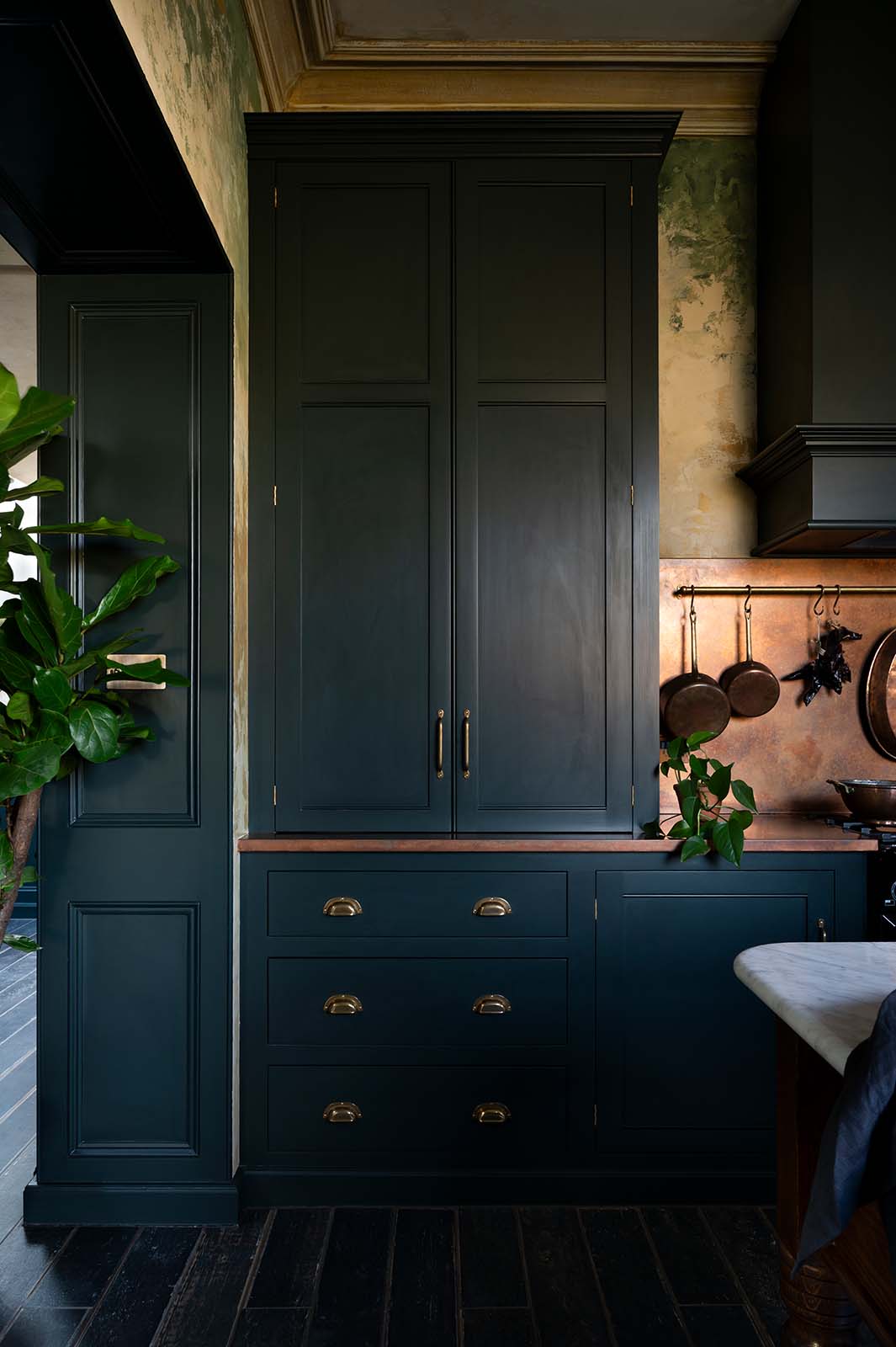 A closer look at an elegant classic kitchen’s cooker run reveals an aged copper worktop, a shaker canopy, counter cabinets, and a marble worktop heirloom dairy table. The pantry-blue counter cabinets stand tall, and beside them is a black gas cooker, copper pans, and fresh chillies hanging from aged brass rails, with a large aged copper splashback behind.