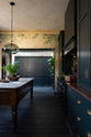 An elegant classic kitchen includes a cooker with an aged copper worktop, a shaker canopy, counter cabinets, and a marble worktop heirloom dairy table. The tall-ceilinged room has a rustic wash finish, with green draping down the walls, resembling overgrown ancient ruins. Beyond the archways leading to the hallway is a large classic English fitted larder, perfect for storage or general pantry use.