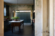 Looking through the archways is an elegant classic kitchen with a cooker run, an aged copper worktop, a Shaker canopy, counter cabinets, and a marble worktop heirloom dairy table. The tall-ceilinged room has a rustic wash finish, with green draping down the walls, resembling overgrown ancient ruins. Beyond the archways leading to the hallway is a large classic English fitted larder, perfect for storage or general pantry use.