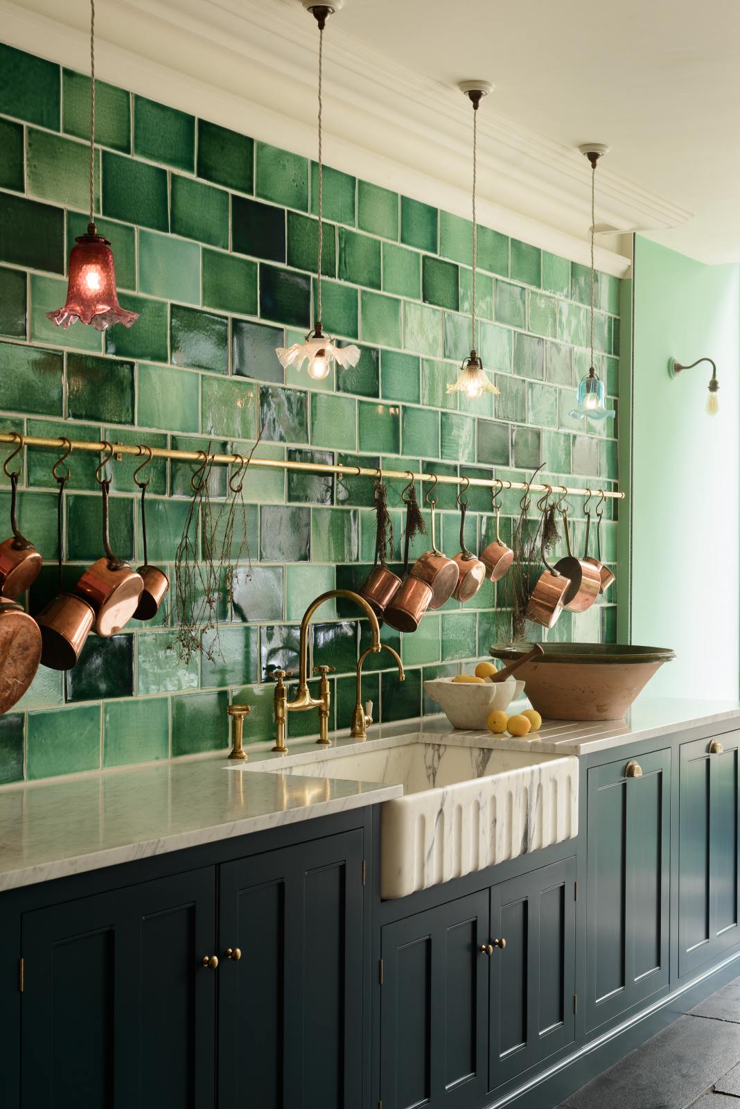 An angled view of a stunning, Bond Street blue kitchen features a sink and cooker run with Arabescato marble worktop, and an aged brass ionian tap. The room features a stunning mix of colours and character, accentuated by elegant antiques such as colourful glass pendant lights, aged brass rails with copper pans, an aged brass pot filler, and deVOL Emerald green London splashback tiles.