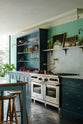 An angled view of a stunning, Bond Street blue kitchen features a cooker run with Arabescato marble worktop and splashback, a bespoke prep table, and a bank of open pantry larder. The room features a stunning mix of colours and character, accentuated by elegant antiques such as a collection of colourful drinking glasses, and an aged brass pot filler.