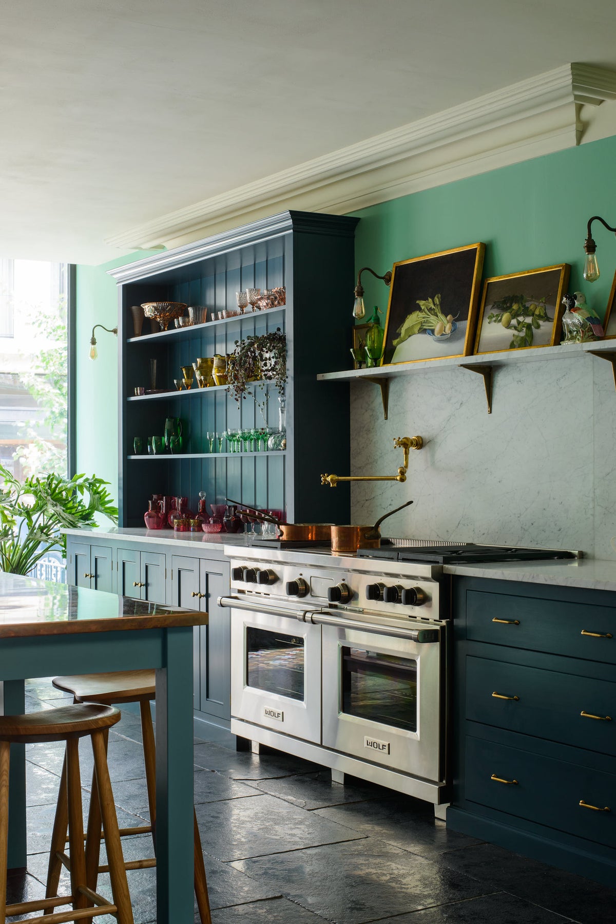 An angled view of a stunning, Bond Street blue kitchen features a cooker run with Arabescato marble worktop and splashback, a bespoke prep table, and a bank of open pantry larder. The room features a stunning mix of colours and character, accentuated by elegant antiques such as a collection of colourful drinking glasses, and an aged brass pot filler.