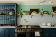 A stunning, Bond Street blue kitchen features a cooker run with Arabescato marble worktop and splashback, and a bank of open pantry larder. The room features a stunning mix of colours and character, accentuated by elegant antiques such as a collection of colourful drinking glasses, and an aged brass pot filler.