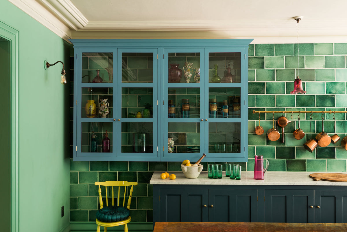 A stunning, Bond Street blue kitchen features a sink run with Arabescato marble worktop, a bespoke prep table, and a large wall cupboard. The room features a stunning mix of colours and character, accentuated by elegant antiques such as artisan ceramics, colourful glass pendant lights, aged brass rails with copper pans, and deVOL Emerald green London splashback tiles.