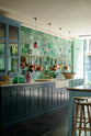 An oblique view of a stunning, Bond Street blue kitchen features a sink run with Arabescato marble worktop, a bespoke prep table, and a large wall cupboard. The room features a stunning mix of colours and character, accentuated by elegant antiques such as artisan ceramics, colourful glass pendant lights, aged brass rails with copper pans, and deVOL Emerald green London splashback tiles.