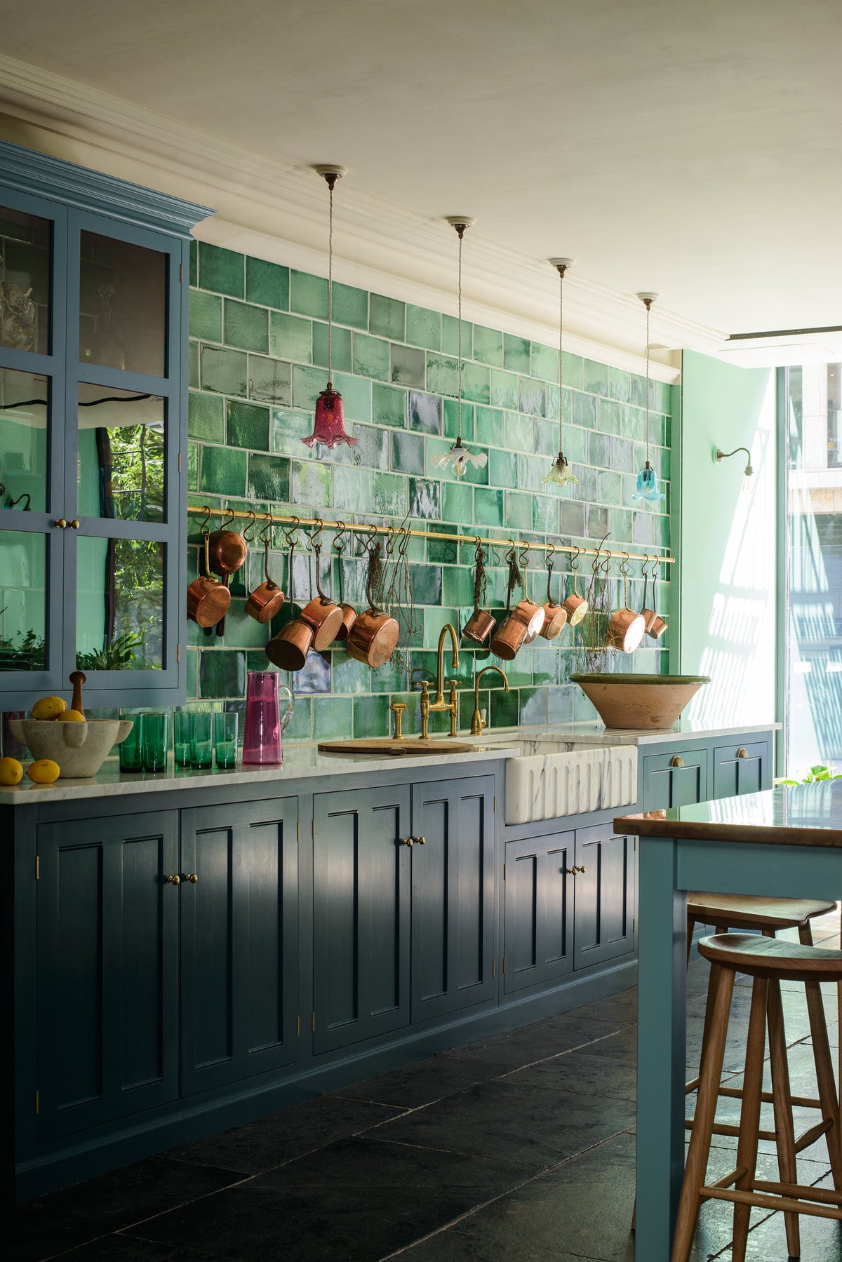 An oblique view of a stunning, Bond Street blue kitchen features a sink run with Arabescato marble worktop, a bespoke prep table, and a large wall cupboard. The room features a stunning mix of colours and character, accentuated by elegant antiques such as artisan ceramics, colourful glass pendant lights, aged brass rails with copper pans, and deVOL Emerald green London splashback tiles.