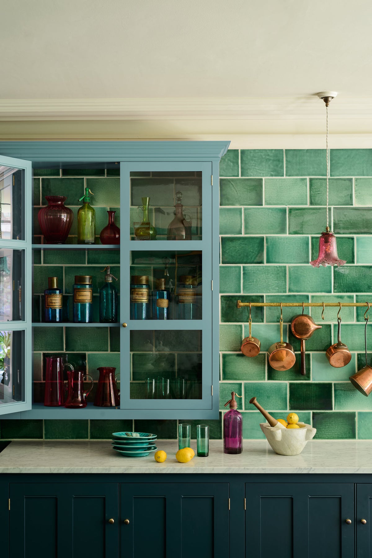 A closer look at a stunning, Bond Street blue kitchen features a sink run with Arabescato marble worktop, and a large wall cupboard. The room features a stunning mix of colours and character, accentuated by elegant antiques such as artisan ceramics, colourful glass pendant lights, aged brass rails with copper pans, and deVOL Emerald green London splashback tiles.