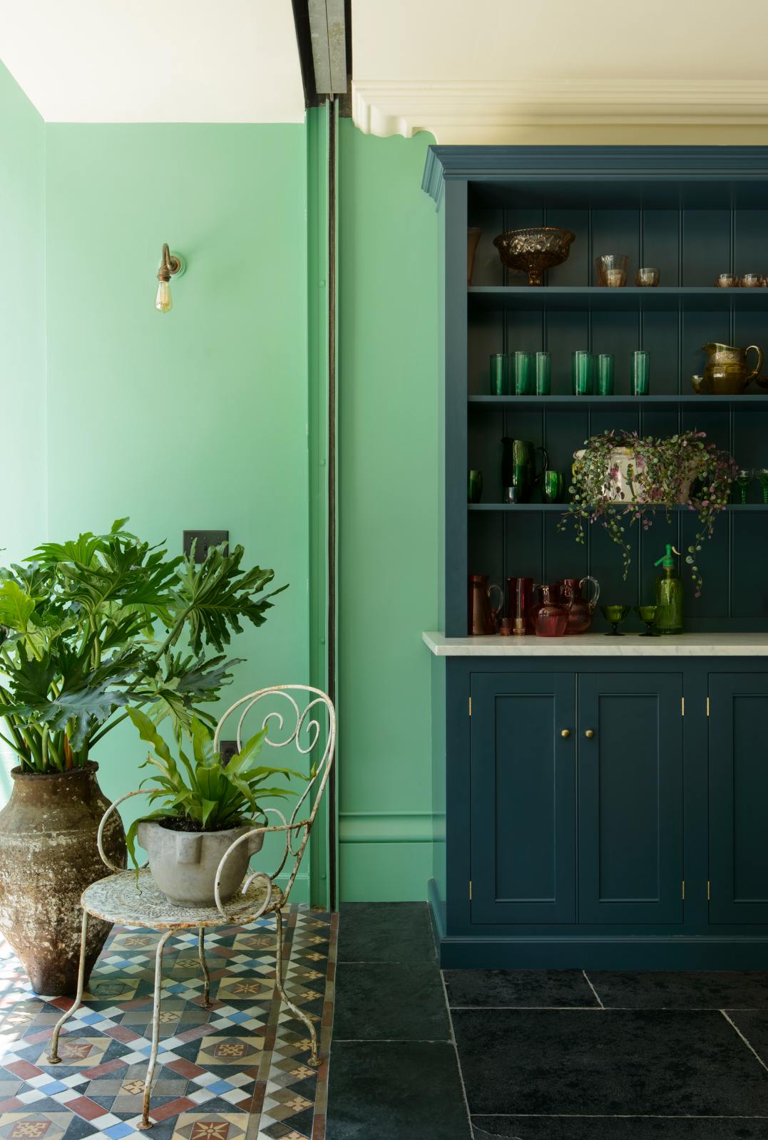 A quiet corner of a stunning, Bond Street blue kitchen features a bank of open pantry larder. The room features a stunning mix of colours and character, accentuated by elegant antiques such as a collection of colourful drinking glasses, potted plants, and artisan ceramics.