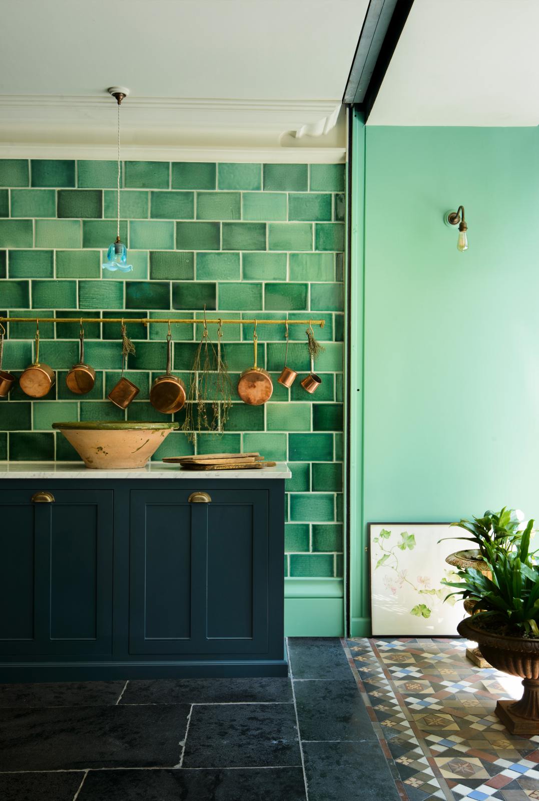 A quiet corner of a stunning, Bond Street blue kitchen features a sink run with Arabescato marble worktop. The room features a stunning mix of colours and character, accentuated by elegant antiques such as artisan ceramics, colourful glass pendant lights, aged brass rails with copper pans, and deVOL Emerald green London splashback tiles.