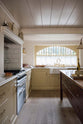 A warm, inviting, muted yellow classic English kitchen features an L-shaped cooker and sink run with a Carrara marble worktop and a large freestanding maroon prep table with a solid oak worktop. The double Belfast sink with an aged brass Ionian tap sits in front of a semi-circular window with an aged brass curtain rail at the bottom half, allowing privacy and ample natural light to brighten the room.