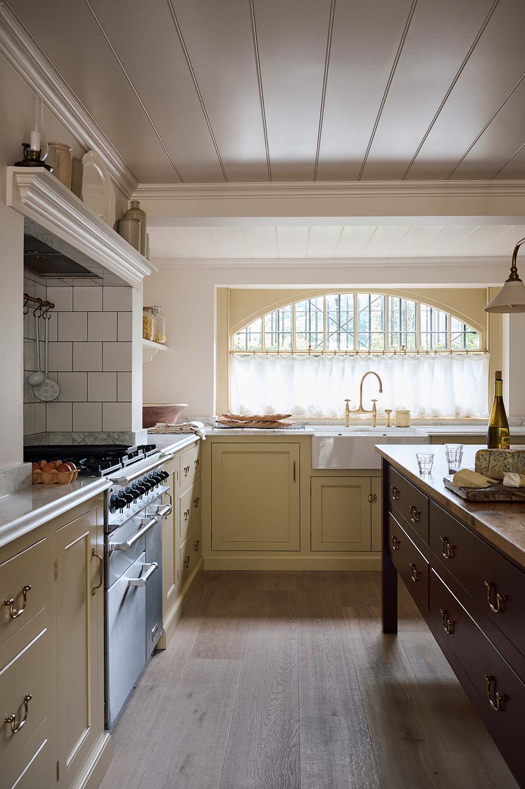 A warm, inviting, muted yellow classic English kitchen features an L-shaped cooker and sink run with a Carrara marble worktop and a large freestanding maroon prep table with a solid oak worktop. The double Belfast sink with an aged brass Ionian tap sits in front of a semi-circular window with an aged brass curtain rail at the bottom half, allowing privacy and ample natural light to brighten the room.