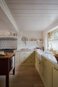 A warm, muted yellow classic English kitchen features an L-shaped area with a Carrara marble worktop for the cooker and sink. A large maroon prep table with a solid oak top adds functionality. The silver gas cooker is surrounded by white porcelain splashback tiles and an aged brass hanging rail. The double Belfast sink, fitted with an aged brass Ionian tap, sits beneath a semi-circular window, with an aged brass curtain rail, allowing for privacy and natural light.