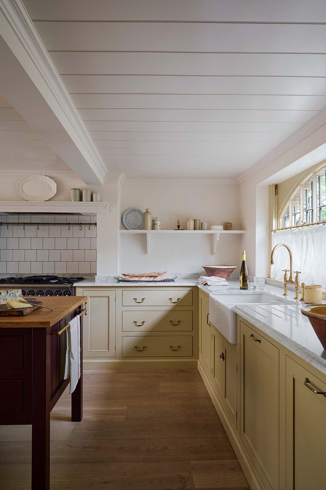 A warm, muted yellow classic English kitchen features an L-shaped area with a Carrara marble worktop for the cooker and sink. A large maroon prep table with a solid oak top adds functionality. The silver gas cooker is surrounded by white porcelain splashback tiles and an aged brass hanging rail. The double Belfast sink, fitted with an aged brass Ionian tap, sits beneath a semi-circular window, with an aged brass curtain rail, allowing for privacy and natural light.