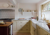 A closer look at the classic muted yellow kitchen reveals an L-shaped configuration with a cooker and sink, complemented by a Carrara marble worktop. A large freestanding dairy table with a solid oak surface adds to the functionality of the space. The sink is positioned beneath a semi-circular window, allowing ample natural light to brighten the room.