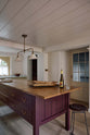 An oblique view of a warm, inviting, muted yellow classic English kitchen features an L-shaped cooker and sink run with a Carrara marble worktop, a large freestanding maroon prep table with a solid oak worktop and a breakfast nook. Adorned with deVOL dark creedy stool, a cherished muted bowl, a deVOL brass heirloom gaselier light, freshly baked baguettes, and wine, this kitchen feels humble and inviting for a family that enjoys cooking.