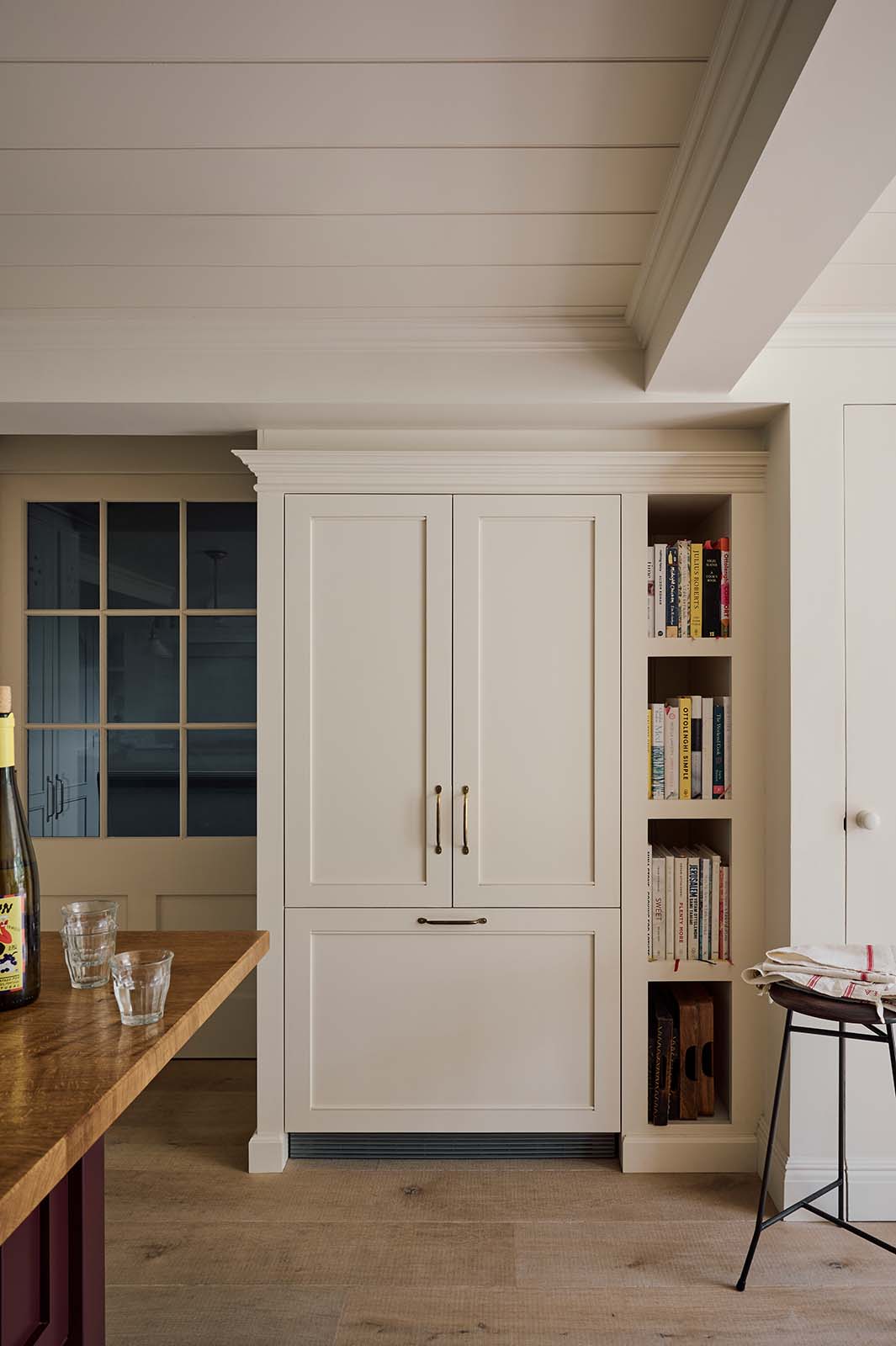 In a quiet corner of a warm, inviting, muted classic English kitchen features a large freestanding maroon prep table with a solid oak worktop and a breakfast nook, a full-height fridge and freezer cupboard and tray slots. Adorned with a deVOL dark creedy stool, books, woods chopping boards, and wine, this kitchen feels humble and inviting for a family that enjoys cooking.