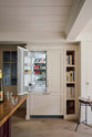 In a quiet corner of a warm, inviting, muted classic English kitchen features a large freestanding maroon prep table with a solid oak worktop and a breakfast nook, a full-height fridge and freezer cupboard and tray slots. Adorned with a deVOL dark creedy stool, books, woods chopping boards, and wine, this kitchen feels humble and inviting for a family that enjoys cooking.