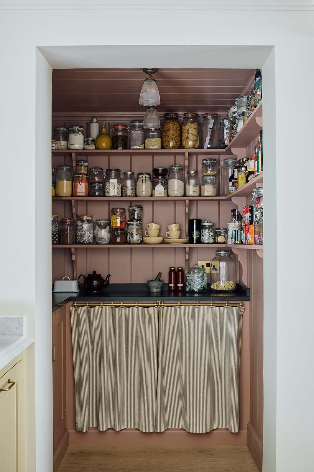 In a warm, inviting, muted yellow classic English kitchen features an walk-in, muted pink pantry with a single door base cabinet, an open base cabinet with an aged brass curtain rail and a black garnite counter, and three top shelves. Adorned with dried ingredients in a glass jards, a muted yellow tea set, a black ceramic tea pot, this kitchen feels humble and inviting for a family that enjoys cooking.