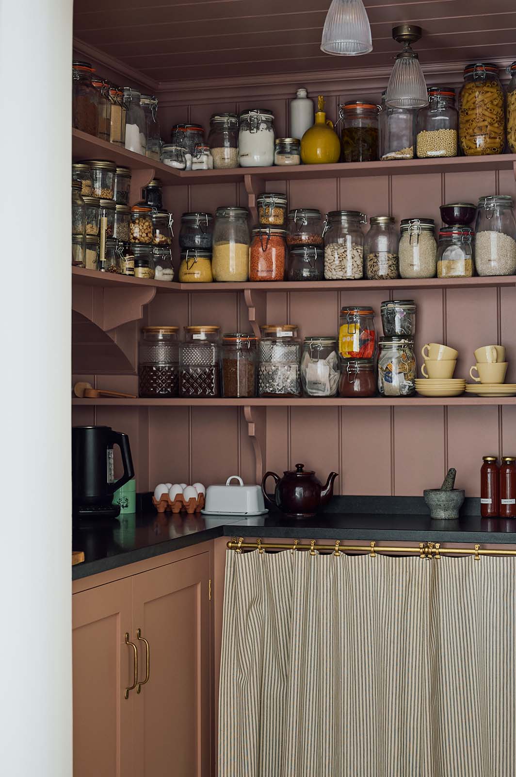 In a warm, inviting, muted yellow classic English kitchen features an walk-in, muted pink pantry with a single door base cabinet, an open base cabinet with an aged brass curtain rail and a black garnite counter, and three top shelves. Adorned with dried ingredients in a glass jards, a muted yellow tea set, a black ceramic tea pot, this kitchen feels humble and inviting for a family that enjoys cooking.
