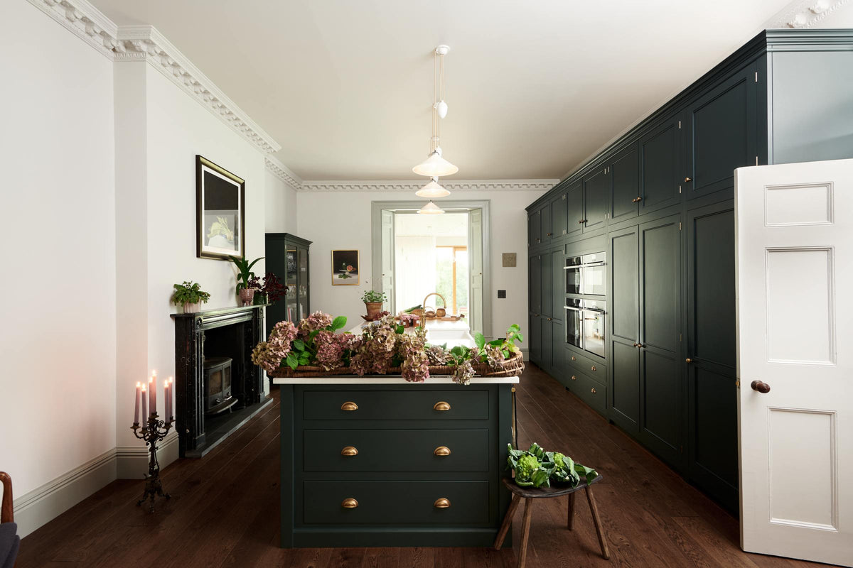 This grand bakehouse green classic English kitchen has large full-height pantry cupboards, oven cabinets, fridge/freezer cupboards, a long kitchen island with a double sink, an aged brass Ionian tap, and a curiosity cupboard. Adorned with white and blush pink hydrangeas, romanesco cauliflower, potted plants, still life paintings, a candelabra, and homemade jams, the room feels homey and welcoming despite its large and decorative appearance.