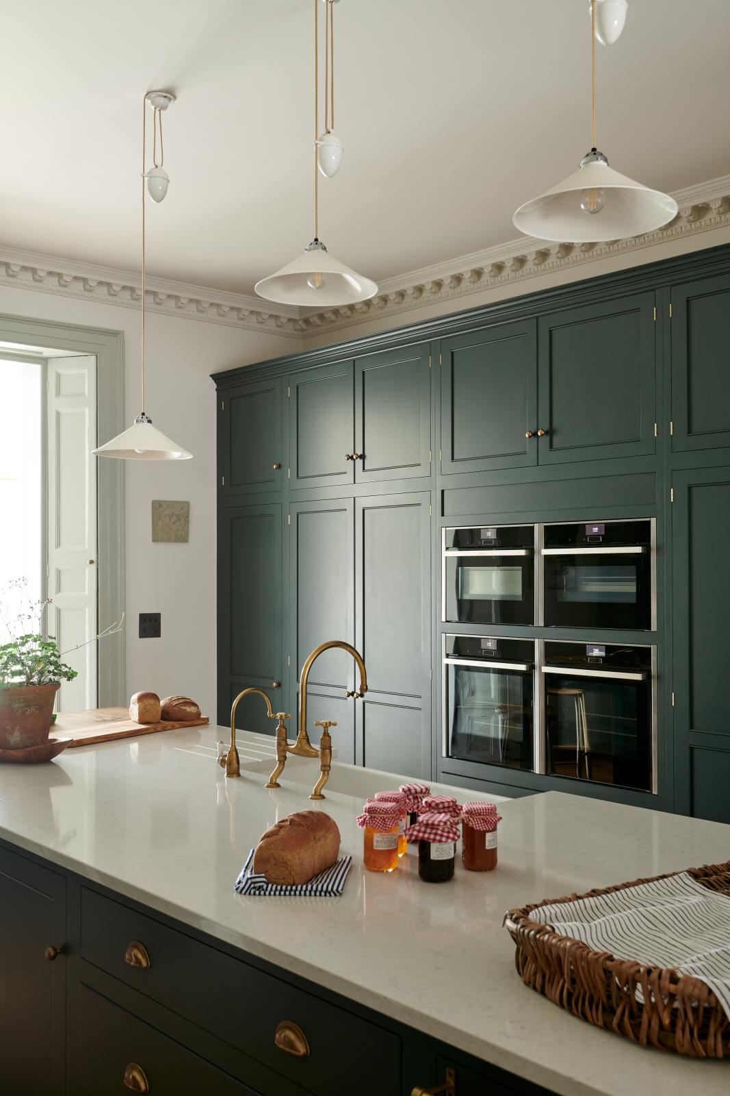 An oblique view of a long kitchen island with a double sink, an aged brass Ionian tap, the large full-height pantry cupboards, oven cabinets and fridge/freezer cupboards at the back of the grand bakehouse green classic English kitchen. Adorned with loaves of bread, a potted plant, still life paintings, and homemade jams, the room feels homey and welcoming despite its large and decorative appearance.