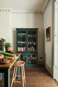 A quiet corner of the grand Bakehouse green classic English kitchen features the curiosity cupboard, a solid oak worktop prep table at the end of the kitchen island with a deVOL bum stool. Adorned with loaves of bread, leeks, red grapes, potted plants, paintings, and books, the room feels homey and welcoming despite its large and decorative appearance.