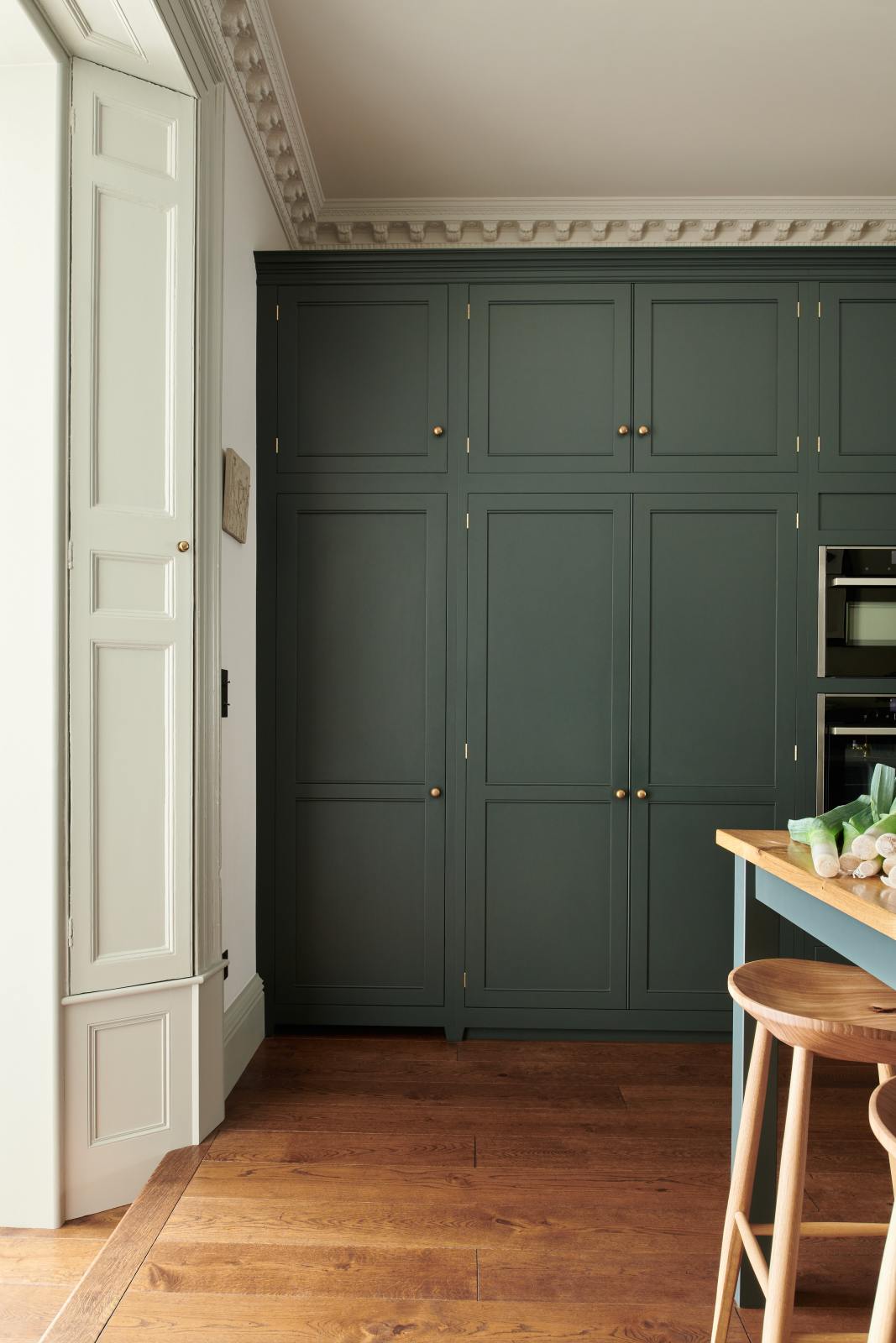 Looking at the edge of a large full-height pantry cupboard, oven cabinets, fridge/freezer cupboard, and a solid oak worktop prep table in the grand bakehouse green classic English kitchen. Adorned with leeks and deVOL wooden bum stools, the room feels homey and welcoming despite its large and decorative appearance.