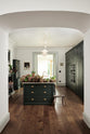 Looking through a large archway is a grand, Bakehouse green classic English kitchen has large full-height pantry cupboards, oven cabinets, fridge/freezer cupboards, a long kitchen island with a double sink, an aged brass Ionian tap, and a curiosity cupboard. Adorned with white and blush pink hydrangeas, romanesco cauliflower, potted plants, still life paintings, a candelabra, and homemade jams, the room feels homey and welcoming despite its large and decorative appearance.