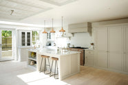 A simple and stylish classic English kitchen features a sink and cooker run, a suspended false chimney breast, a wall cupboard, a glazed larder, and a kitchen island topped with Carrara marble. Beautifully decorated with light copper pendant lights, a silver toaster, a silver kettle, muted ceramics, and white splashback tiles, it creates a functional and beautiful kitchen space.