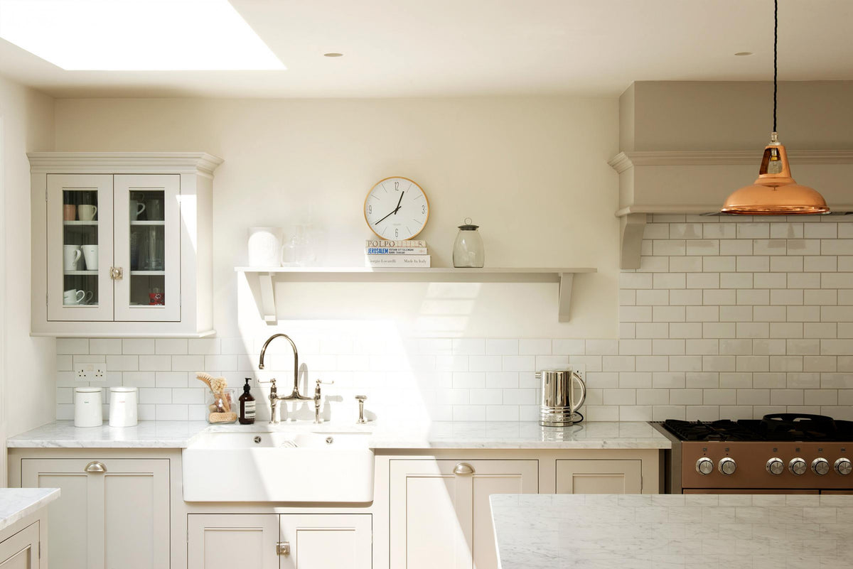 This close-up of a simple and stylish classic English kitchen highlights the sink and cooker run, featuring a double Belfast sink, an antique silver Ionian tap, and a Carrara marble worktop. It includes a glazed wall cupboard and a shelf. Beautifully decorated with a light copper pendant light, glass containers, white ceramics, a silver kettle, wooden cleaning equipment, and white splashback tiles, this space creates a functional and beautiful kitchen atmosphere.