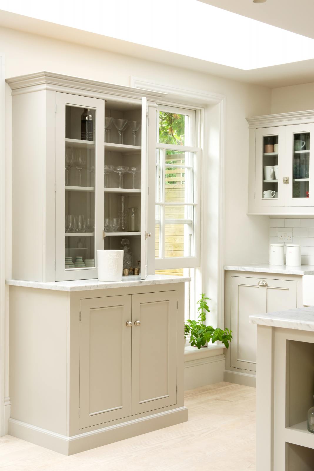 A close-up of a simple and stylish classic English kitchen’s glazed larder with a Carrara marble worktop. Beautifully decorated with white ceramics, drinking glasses, a basil plant, and white splashback tiles, it creates a functional and beautiful kitchen space.