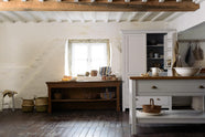 A beautiful white linen kitchen features a freestanding table, a pantry cupboard, and a collection of vintage furniture and items, reflecting on the well-reserved historical space with its original plastered walls, wooden beams, and oak floors.
