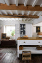 A beautiful white linen kitchen features a freestanding table, an open pantry cupboard, and a collection of vintage furniture and items, reflecting on the well-reserved historical space with its original plastered walls, wooden beams, and oak floors.