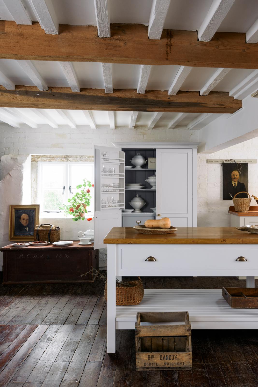 A beautiful white linen kitchen features a freestanding table, an open pantry cupboard, and a collection of vintage furniture and items, reflecting on the well-reserved historical space with its original plastered walls, wooden beams, and oak floors.