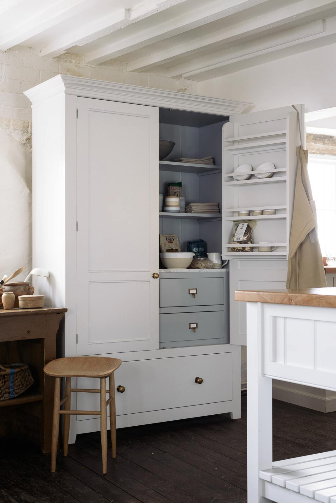A beautiful linen white kitchen features an open pantry cupboard with a spice rack, marble counter, shelves and closed drawers, and a collection of vintage furniture and items reflecting on the well-reserved historical space with its original plastered walls, wooden beams and oak floors.