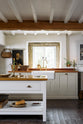 An angled view of a beautiful white linen kitchen featuring a sink run with aged brass Ionian taps, a freestanding table, and a collection of vintage furniture and items reflecting on the well-reserved historical space with its original plastered walls, wooden beams and oak floors.