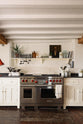 A beautiful white linen kitchen features a cooker run, a silver gas stove, a shaker shelf, and a collection of vintage furniture and items reflecting on the well-reserved historical space with its original plastered walls, wooden beams and oak floors.