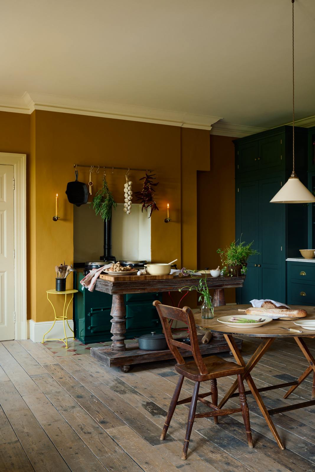 An angled view of a lovely modern kitchen that retains the charm of its original structure and flooring, using the three primary colours in a muted, mature palette. The spacious kitchen features a Clerkenwell Blue full-height pantry cabinet and a long sink run with a marble worktop. An emerald green gas stove sits within the chimney breast, accompanied by a deVOL cream pendant light above a vintage round wooden table, with matching chairs and a prep table.