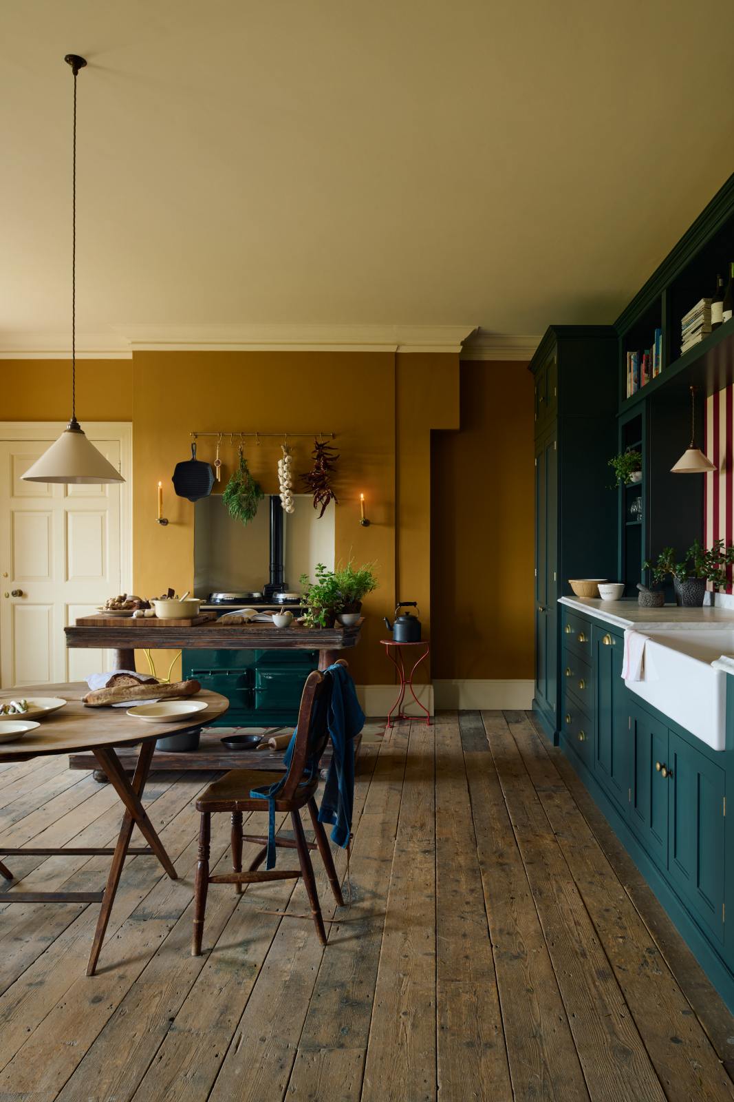 A lovely modern kitchen that retains the charm of its original structure and flooring, using the three primary colours in a muted, mature palette. The spacious kitchen features a Clerkenwell Blue full-height pantry cabinet, a long sink run with a marble worktop, and an open countertop cabinet and shelf. An emerald green stove sits within the chimney breast, accompanied by a deVOL cream pendant light above a vintage round wooden table, with matching chairs and a prep table.